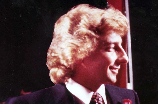 Barry Manilow in profile in a dark suit with a red rose on the lapel, photographed in November 1979, four years after Mandy