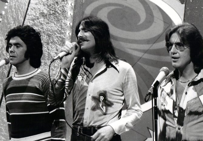Three Dog Night singers Danny Hutton, Chuck Negron and Cory Wells at microphones on an NBC television stage in 1975, the voices behind the Three Dog Night songs of the 70s